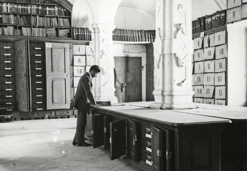 Schwarzweißbild eines Mannes, der über einen Tisch gebeugt in einem großen Archivraum mit Gewölbedecke steht.